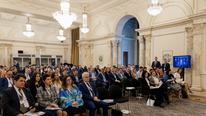 Konferences zāle ar augstiem griestiem un lustru apgaismojumu, kur rindās sēž pasākuma dalībnieki, klausoties prezentāciju; zāles labajā pusē redzams ekrāns ar vizuālo materiālu.