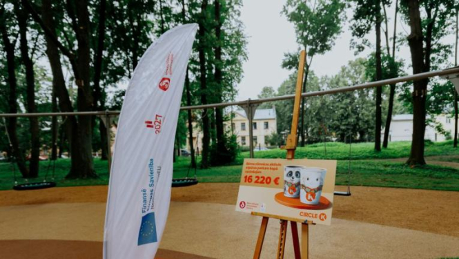 Āra teritorija ar rotaļu laukumu fonā. Priekšplānā redzama balta karogs ar tekstu un logotipiem, kā arī molberts ar plakātu, uz kura attēloti divi bundžu attēli un teksts: "Bērnu pilsētas aktivitāšu centra izveidei piešķirts līdzfinansējums 16 220 €".