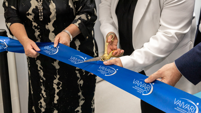 Trīs cilvēki piedalās lentes griešanas ceremonijā pie VAIVARI Nacionālā rehabilitācijas centra. Viens no viņiem ar lielām zelta šķērēm pārgriež zilu lenti ar centra nosaukumu, atzīmējot jaunas ēkas vai pakalpojuma atklāšanu.