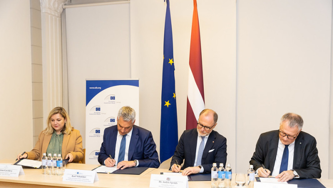 4 pārstāvji paraksta dokumentus pie galda, fonā redzamas Eiropas Savienības un Latvijas karogi, kā arī Eiropas Investīciju bankas (EIB) baneris ar vietni www.eib.org. Attēls no oficiālas parakstīšanas ceremonijas.