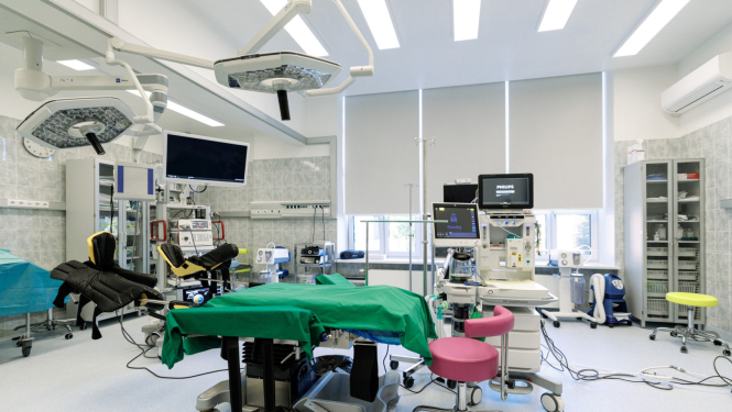 Mūsdienīga operāciju zāle ar centrāli novietotu operāciju galdu, kas pārklāts ar zaļu pārklāju. Telpā redzama medicīniskā aparatūra – ķirurģiskās lampas, monitori, anestēzijas iekārta un citi instrumenti. Fonā redzami plaukti un skapji ar medicīnas piederumiem, kā arī logi ar daļēji aizvilktām žalūzijām. Priekšplānā – rozā krēsls.