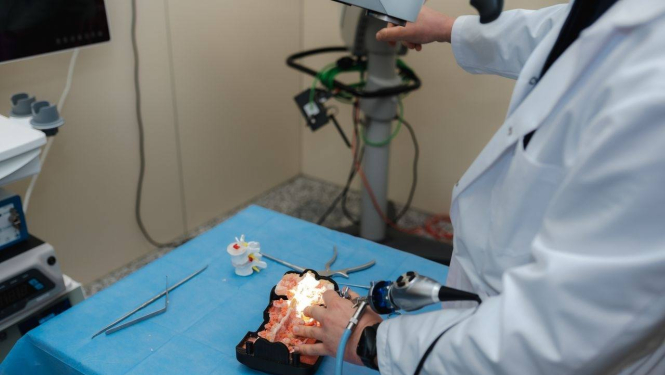 Persona baltā halātā veic manipulācijas ar medicīnisku vai anatomisku galvas/galvas smadzeņu modeli, kas novietots uz zilas ķirurģiskās drānas. Tiek izmantoti medicīnas instrumenti, tostarp endoskops vai tam līdzīga ierīce. Apkārt redzami dažādi rīki un aprīkojums, norādot uz medicīnas laboratoriju vai apmācību telpu, kur notiek praktiska procedūra vai mācību sesija.