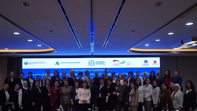 Grupas foto no starptautiskās konferences par dzimumlīdztiesību valsts pārvaldē Taškentā; dalībnieki pozē kopā konferenču zālē, fonā redzams liels ekrāns ar konferences nosaukumu un partneru logotipiem.