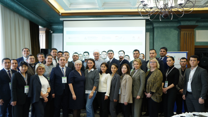 Liela profesionāļu grupa formālā apģērbā pozē kopbildē konferenču zālē; fonā redzams projektora ekrāns, lustras un pasākuma baneri.