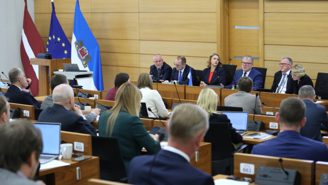 Attēlā redzama sanāksme, kurā piedalās vairāki cilvēki, sēžot pie galda un klausoties prezentāciju vai diskusiju. Priekšplānā ir vairāki cilvēki ar klēpjdatoriem un dokumentiem. Fonā ir Latvijas karogs, Eiropas Savienības karogs un vēl viens karogs ar ģerboni.