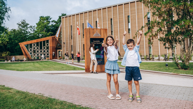 Divi bērni stāv pie mūsdienīgas ēkas ar koka fasādi un vertikāliem logiem, priekšā redzamas karogu mastas un uzraksts 'Institute of Physics Research', apkārt cilvēki uz zāliena un bruģētiem celiņiem.