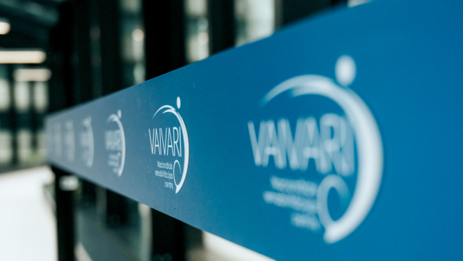 Zila stikla vai sienas virsma ar atkārtotu baltu “VAVARI” logotipu, fotografēta perspektīvā garā gaitenī.