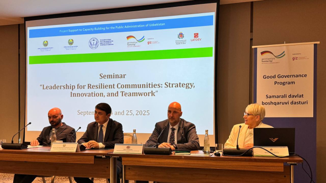 Seminārs “Līderība noturīgām kopienām: stratēģija, inovācijas un komandas darbs”, kas notika 2025. gada 24. un 25. septembrī. Attēlā redzami četri cilvēki, kas sēž pie garas galda ar mikrofonu priekšā. Fonā redzams liels ekrāns ar semināra nosaukumu, datumu un organizāciju logotipiem. Labajā pusē novietots plakāts ar uzrakstu “Good Governance Program” angļu un citā valodā. Telpa ir konferenču zāle ar paklāju un profesionālu iekārtojumu