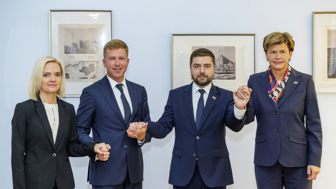 Četri cilvēki formālā biznesa apģērbā stāv rindā, sadevušies rokās, oficiāla pasākuma laikā pie sienas ar ierāmētiem attēliem fonā.