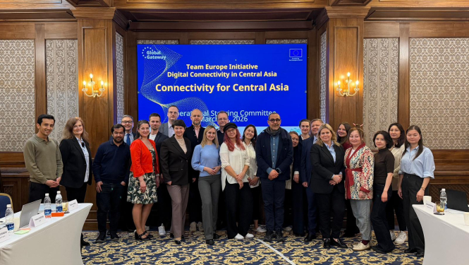 Grupas foto pie sanāksmju galda klasiskā konferenču telpā. Fonā redzams liels ekrāns ar tekstu “Team Europe Initiative – Digital Connectivity in Central Asia. Connectivity for Central Asia. Operational Steering Committee. March 2026”. Cilvēki stāv rindā ekrāna priekšā, telpā redzamas koka sienas, lampas un darba galdi ar dokumentiem.