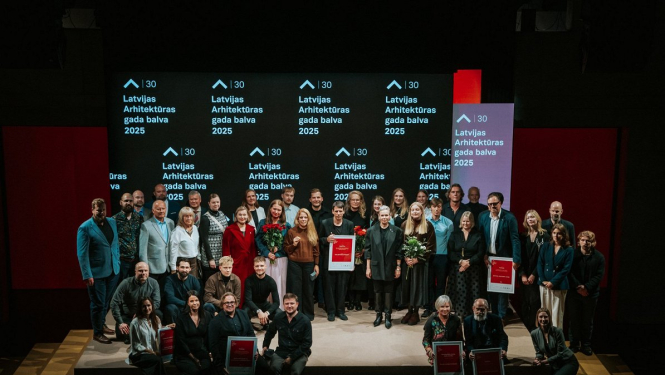 Grupas fotogrāfija ar cilvēkiem, kas stāv uz skatuves un tur rokās balvas. Fonā redzams ekrāns ar uzrakstu "Latvijas Arhitektūras gada balva 2025"