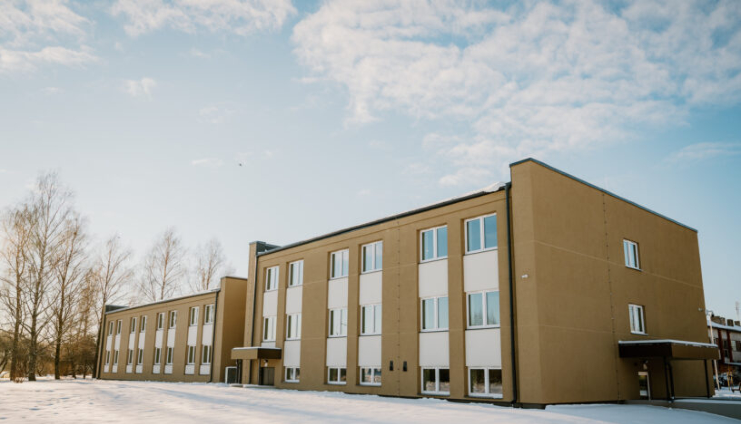 Divstāvu ēka ar gaiši brūnu fasādi un baltām logu ailēm, redzama ziemas ainavā ar sniega klātu zemi priekšplānā. Ēkai ir plakans jumts, vairākas rindas logu, un pie ieejas ir neliels jumtiņš. Fonā redzami kaili koki un gaišas debesis ar dažiem mākoņiem.
