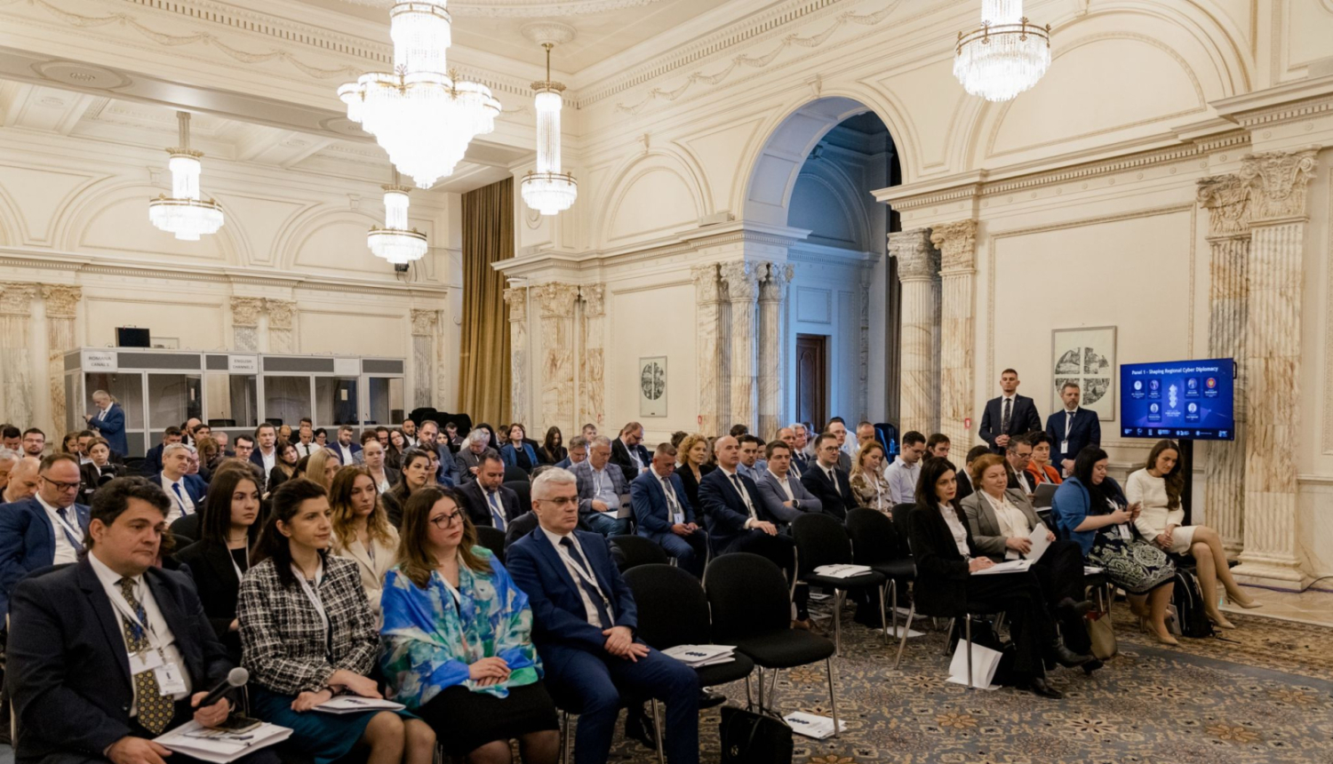 Konferences zāle ar augstiem griestiem un lustru apgaismojumu, kur rindās sēž pasākuma dalībnieki, klausoties prezentāciju; zāles labajā pusē redzams ekrāns ar vizuālo materiālu.