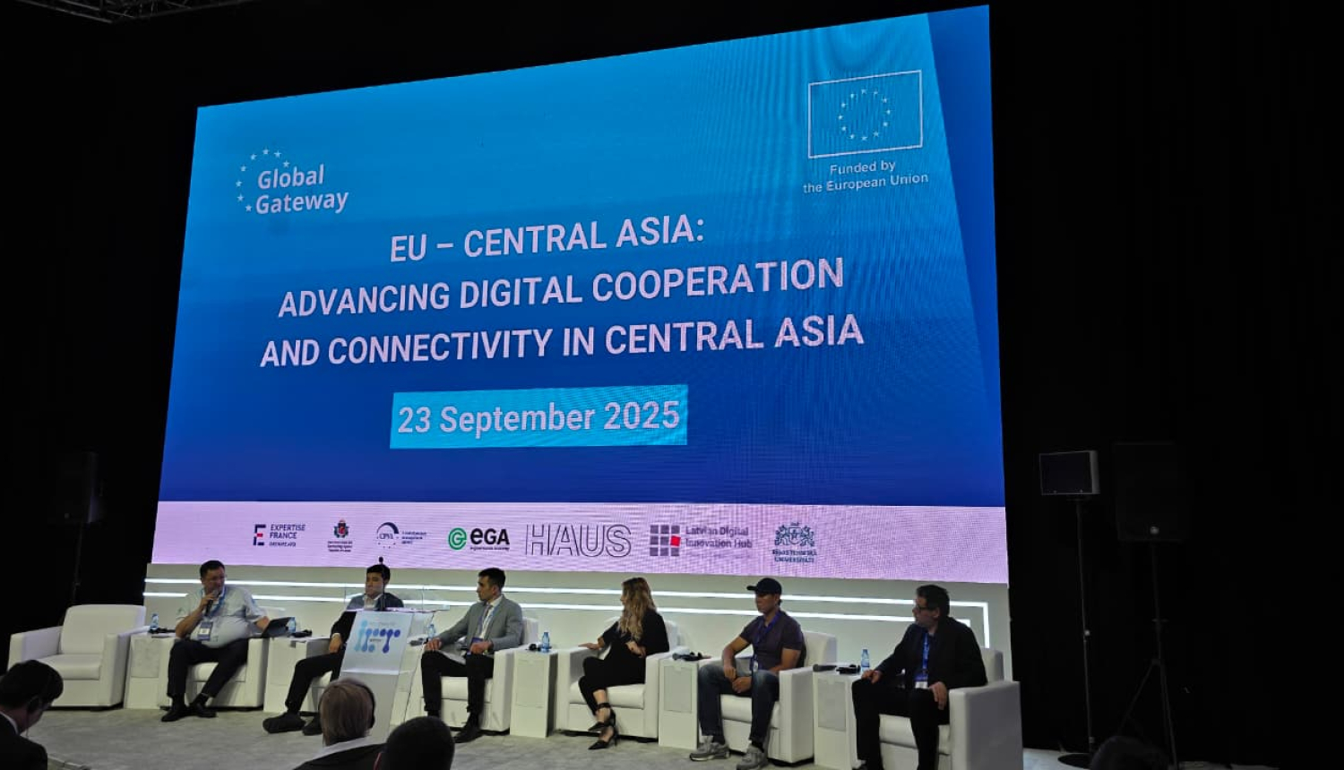 Konferences paneļdiskusija ar pieciem runātājiem uz skatuves, fonā liels ekrāns ar pasākuma nosaukumu “EU - Central Asia: Advancing Digital Cooperation and Connectivity in Central Asia” un datumu “2025. gada 23. septembris”. Pasākums notiek Eiropas Savienības finansētās iniciatīvas “Global Gateway” ietvaros. Ekrāna apakšā redzami dažādu organizāciju logotipi