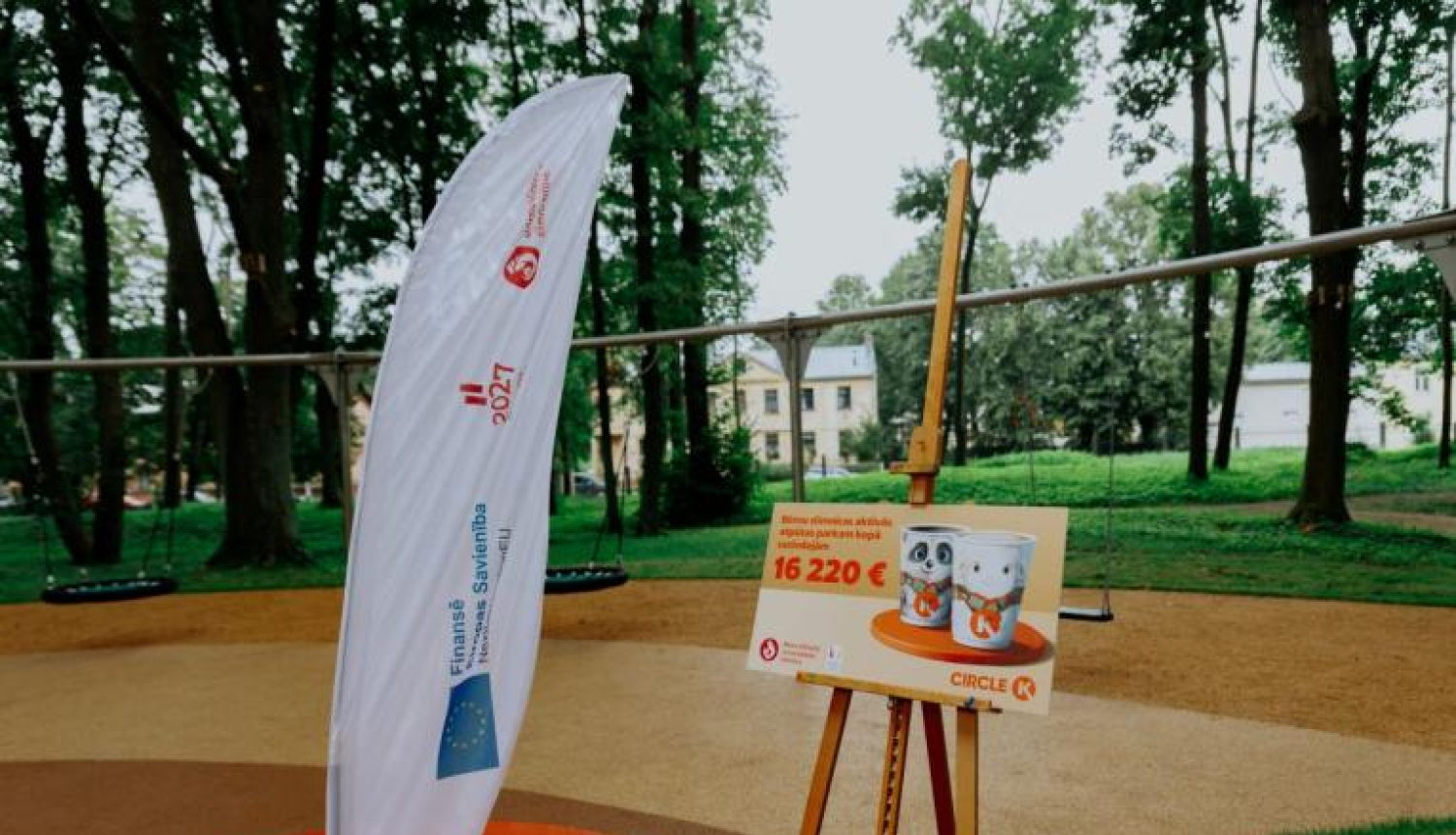 Āra teritorija ar rotaļu laukumu fonā. Priekšplānā redzama balta karogs ar tekstu un logotipiem, kā arī molberts ar plakātu, uz kura attēloti divi bundžu attēli un teksts: "Bērnu pilsētas aktivitāšu centra izveidei piešķirts līdzfinansējums 16 220 €".