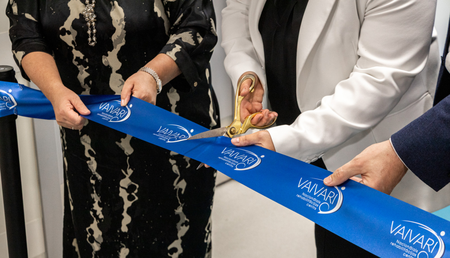 Trīs cilvēki piedalās lentes griešanas ceremonijā pie VAIVARI Nacionālā rehabilitācijas centra. Viens no viņiem ar lielām zelta šķērēm pārgriež zilu lenti ar centra nosaukumu, atzīmējot jaunas ēkas vai pakalpojuma atklāšanu.