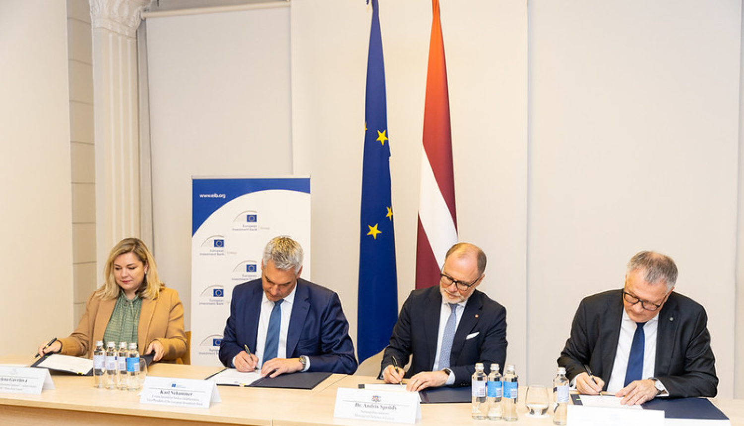 4 pārstāvji paraksta dokumentus pie galda, fonā redzamas Eiropas Savienības un Latvijas karogi, kā arī Eiropas Investīciju bankas (EIB) baneris ar vietni www.eib.org. Attēls no oficiālas parakstīšanas ceremonijas.