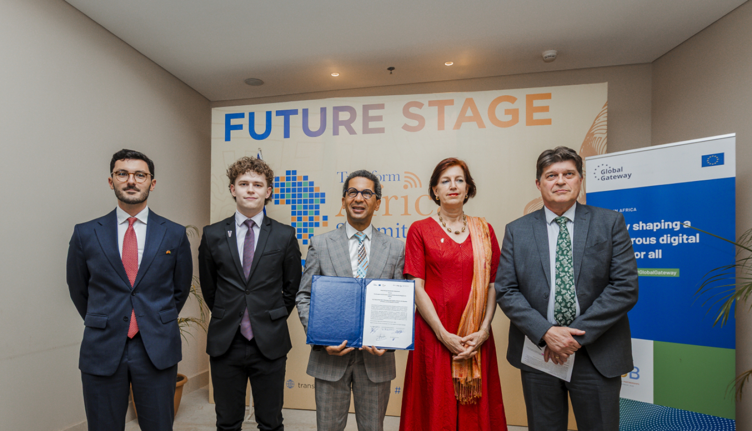 Pieci cilvēki stāv kopā pie sienas ar uzrakstu “Future Stage” un “Transform Africa Summit 2025”, viens tur atvērtu dokumentu mapē. Labajā pusē redzams plakāts ar Eiropas Savienības logotipu un tekstu par digitālās nākotnes veidošanu