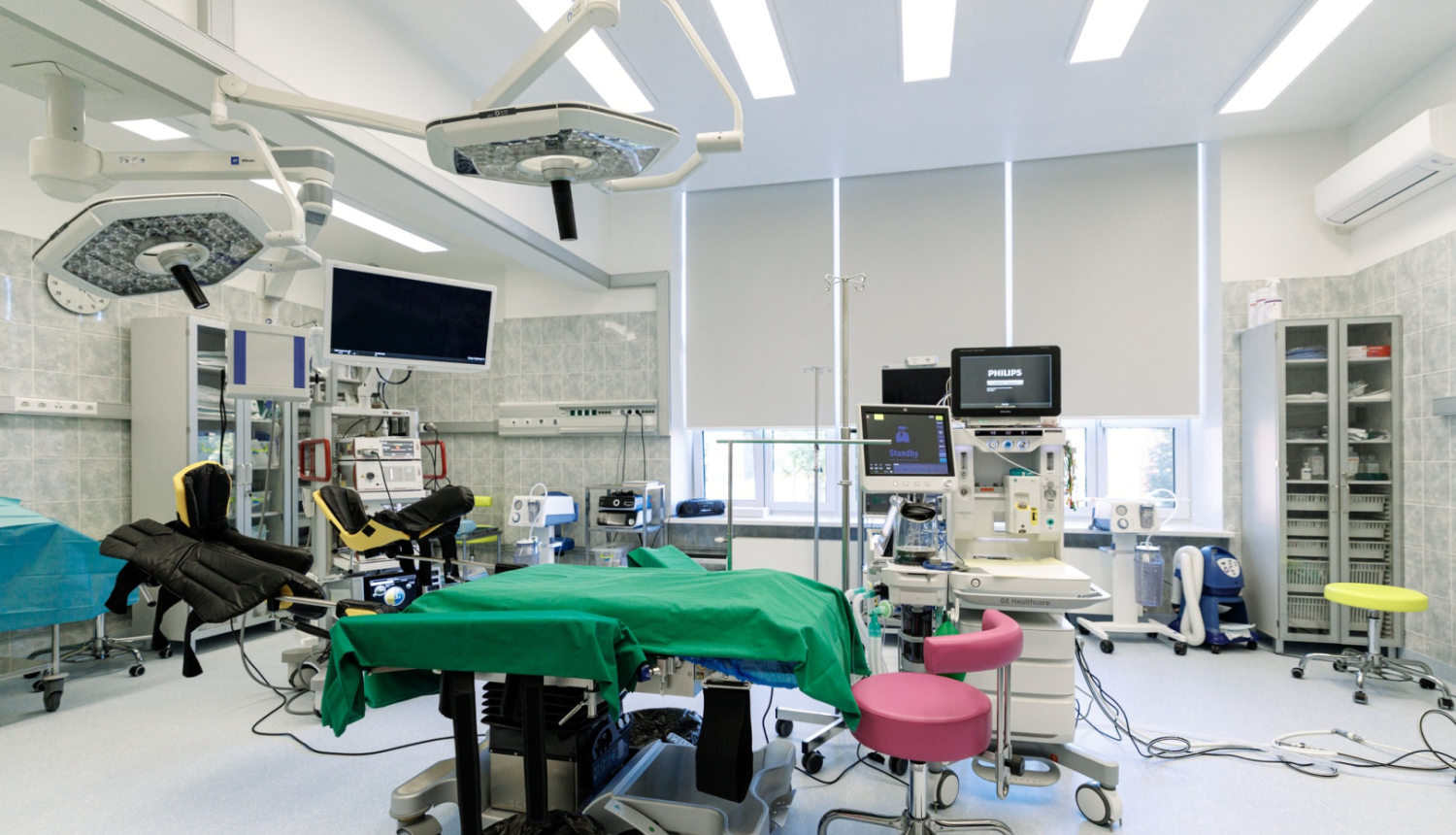 Mūsdienīga operāciju zāle ar centrāli novietotu operāciju galdu, kas pārklāts ar zaļu pārklāju. Telpā redzama medicīniskā aparatūra – ķirurģiskās lampas, monitori, anestēzijas iekārta un citi instrumenti. Fonā redzami plaukti un skapji ar medicīnas piederumiem, kā arī logi ar daļēji aizvilktām žalūzijām. Priekšplānā – rozā krēsls.