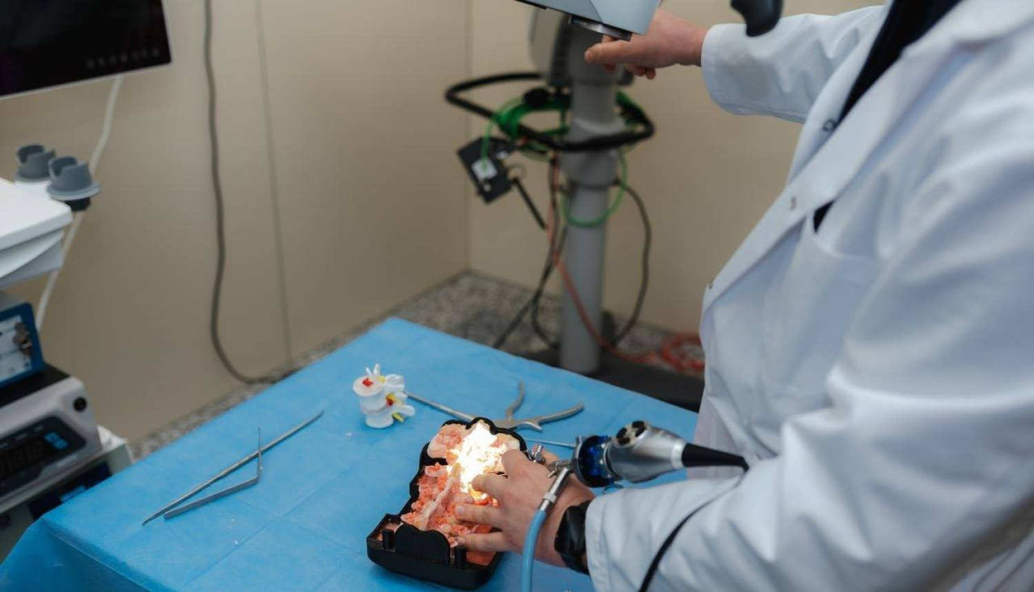 Persona baltā halātā veic manipulācijas ar medicīnisku vai anatomisku galvas/galvas smadzeņu modeli, kas novietots uz zilas ķirurģiskās drānas. Tiek izmantoti medicīnas instrumenti, tostarp endoskops vai tam līdzīga ierīce. Apkārt redzami dažādi rīki un aprīkojums, norādot uz medicīnas laboratoriju vai apmācību telpu, kur notiek praktiska procedūra vai mācību sesija.