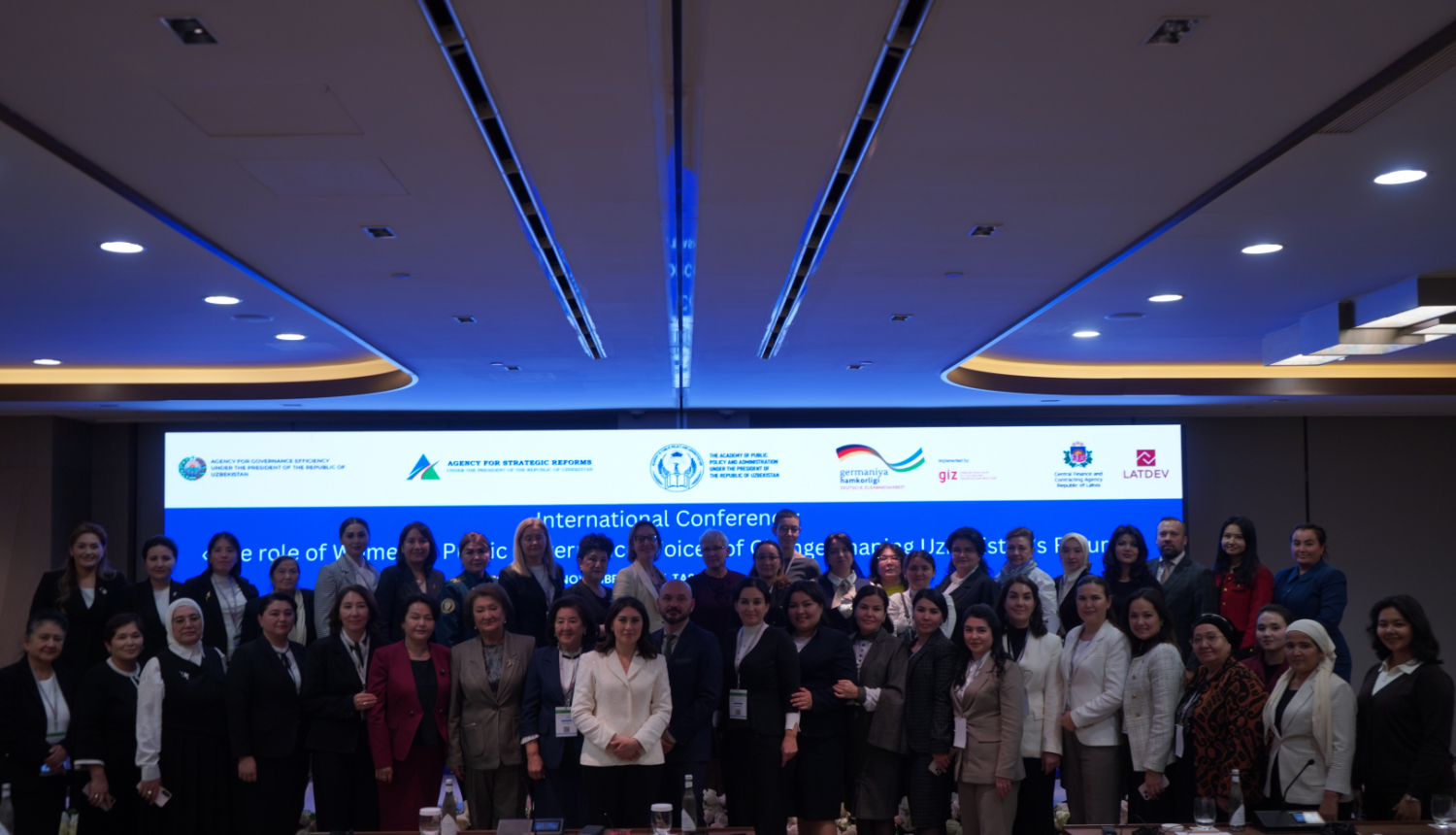 Grupas foto no starptautiskās konferences par dzimumlīdztiesību valsts pārvaldē Taškentā; dalībnieki pozē kopā konferenču zālē, fonā redzams liels ekrāns ar konferences nosaukumu un partneru logotipiem.