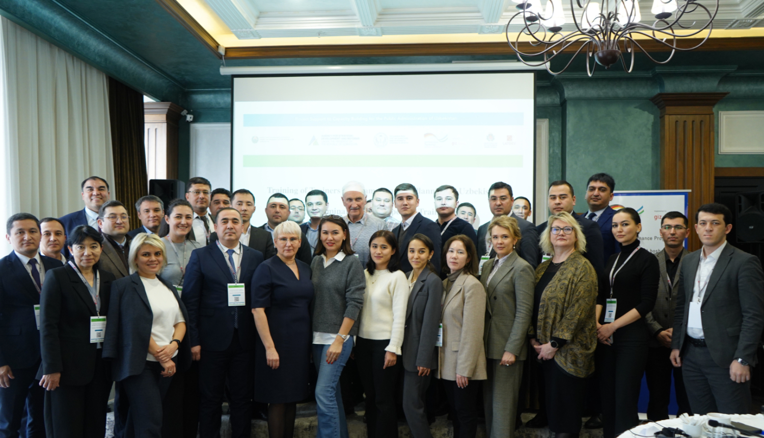 Liela profesionāļu grupa formālā apģērbā pozē kopbildē konferenču zālē; fonā redzams projektora ekrāns, lustras un pasākuma baneri.