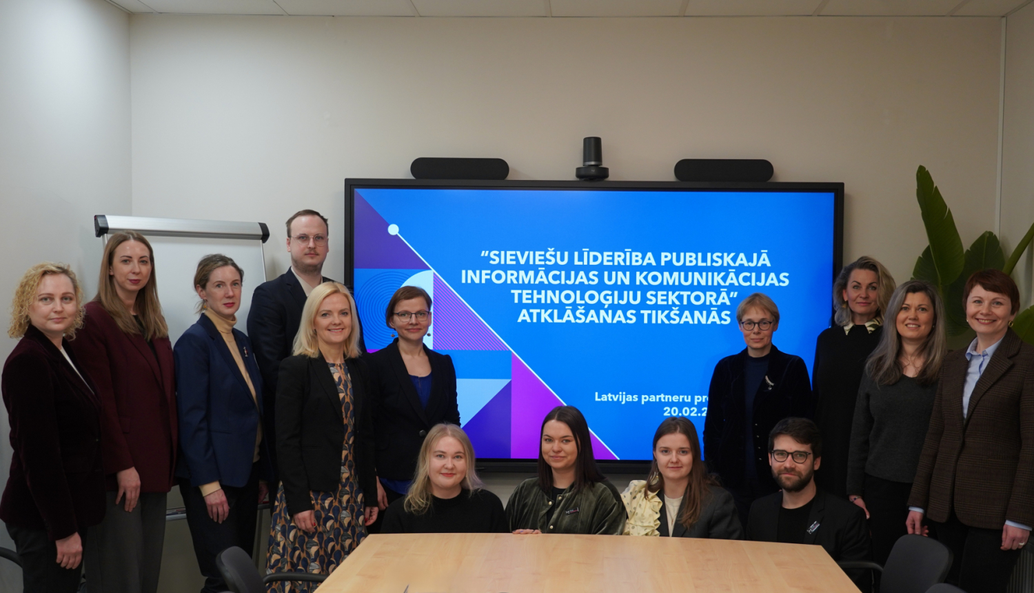 Grupas foto sanāksmē pie prezentāciju ekrāna, uz kura redzams pasākuma nosaukums “Sieviešu līderība publiskajā informācijas un komunikācijas tehnoloģiju sektorā” atklāšanas tikšanās. Dalībnieki nostājušies rindā ekrāna priekšā, fonā redzama tāfele un telpas interjers