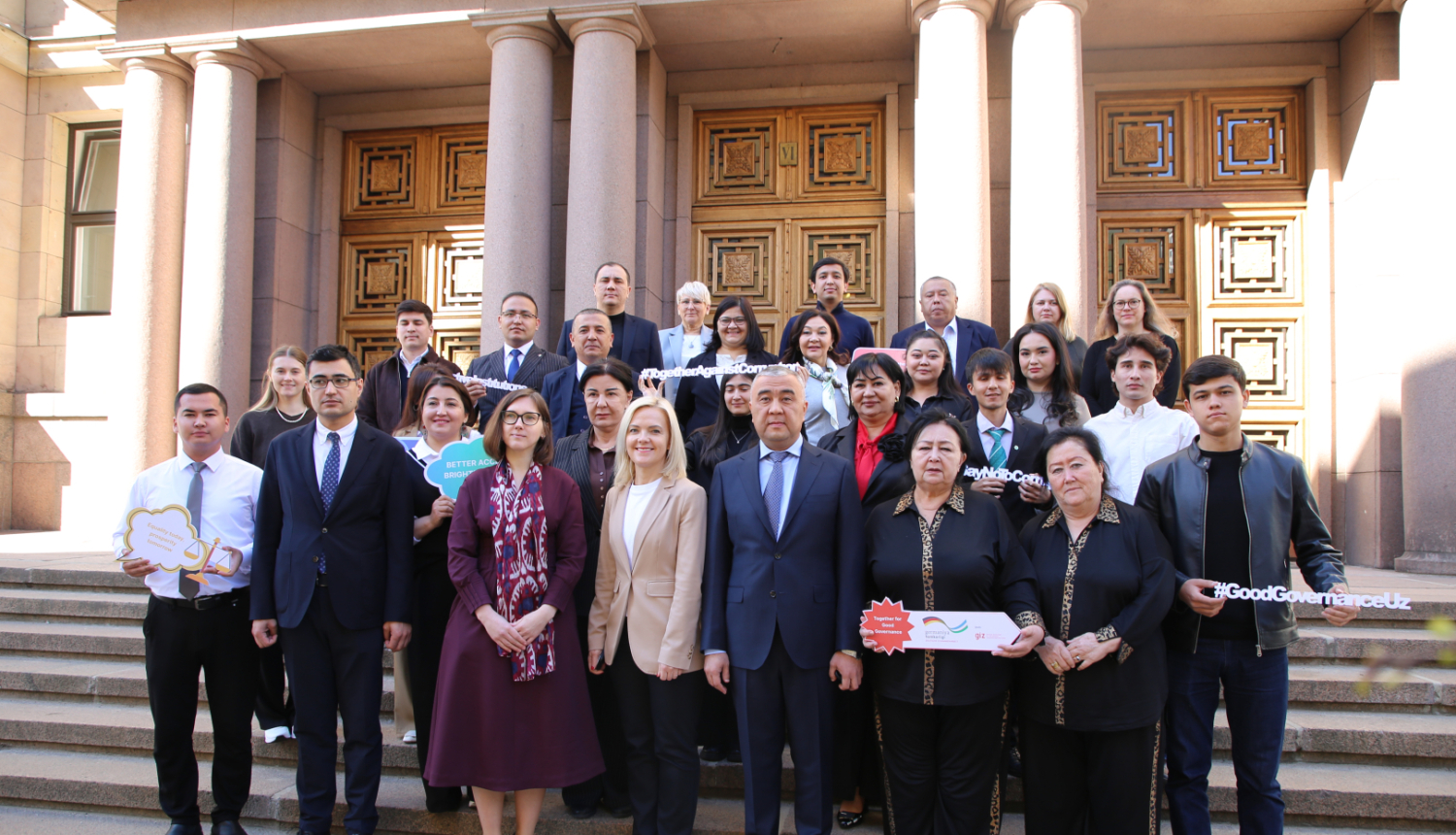 Grupas foto ar vairāk nekā 20 cilvēkiem, kas stāv uz kāpnēm pie monumentālas ēkas ar kolonnām un ornamentētām durvīm. 