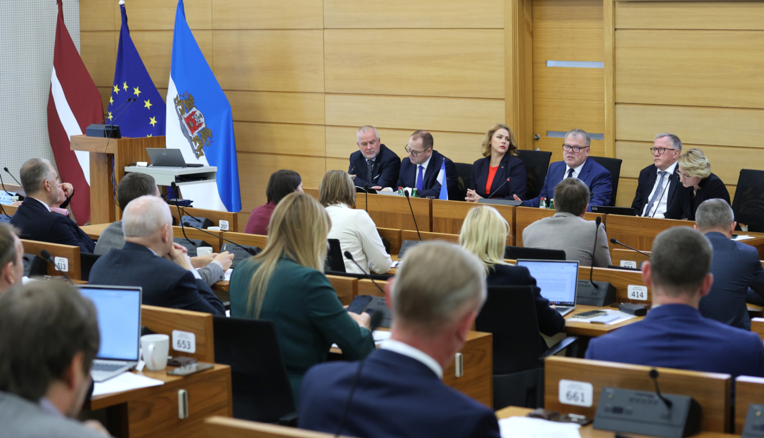 Attēlā redzama sanāksme, kurā piedalās vairāki cilvēki, sēžot pie galda un klausoties prezentāciju vai diskusiju. Priekšplānā ir vairāki cilvēki ar klēpjdatoriem un dokumentiem. Fonā ir Latvijas karogs, Eiropas Savienības karogs un vēl viens karogs ar ģerboni.