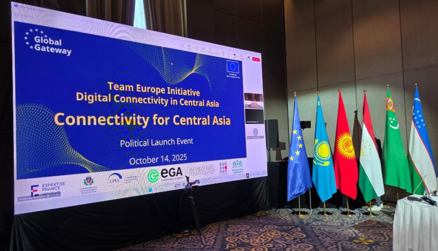 Konferences telpa ar lielu ekrānu, uz kura redzams uzraksts: ‘Global Gateway – Team Europe Initiative – Digital Connectivity in Central Asia – Political Launch Event – October 14, 2025’. Zem teksta izvietoti organizāciju logotipi, tostarp Expertise France, EGA un HAUS. Labajā pusē redzami vairāku valstu karogi: Eiropas Savienības, Kazahstānas, Kirgizstānas, Tadžikistānas, Turkmenistānas un Uzbekistānas