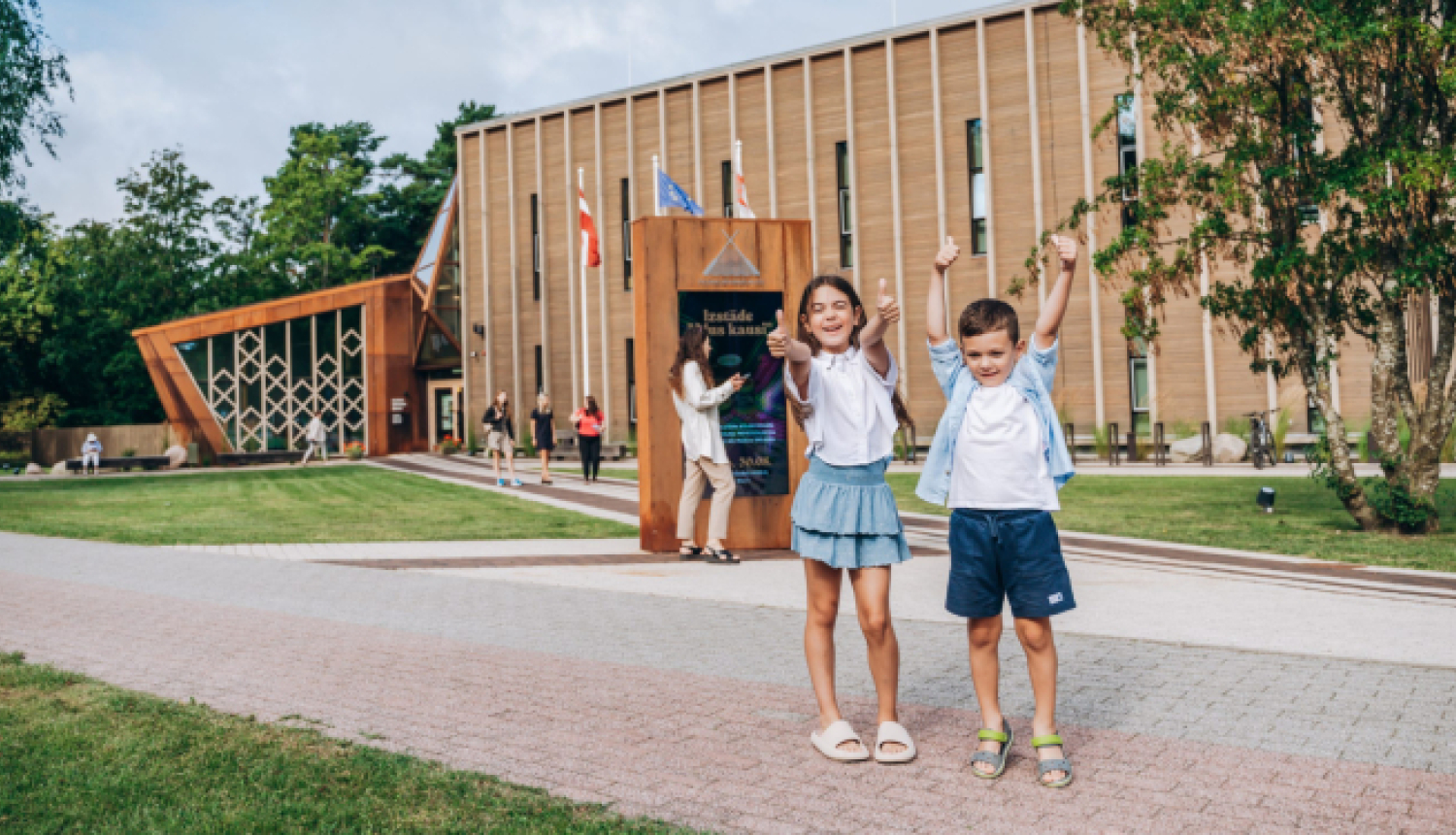 Divi bērni stāv pie mūsdienīgas ēkas ar koka fasādi un vertikāliem logiem, priekšā redzamas karogu mastas un uzraksts 'Institute of Physics Research', apkārt cilvēki uz zāliena un bruģētiem celiņiem.