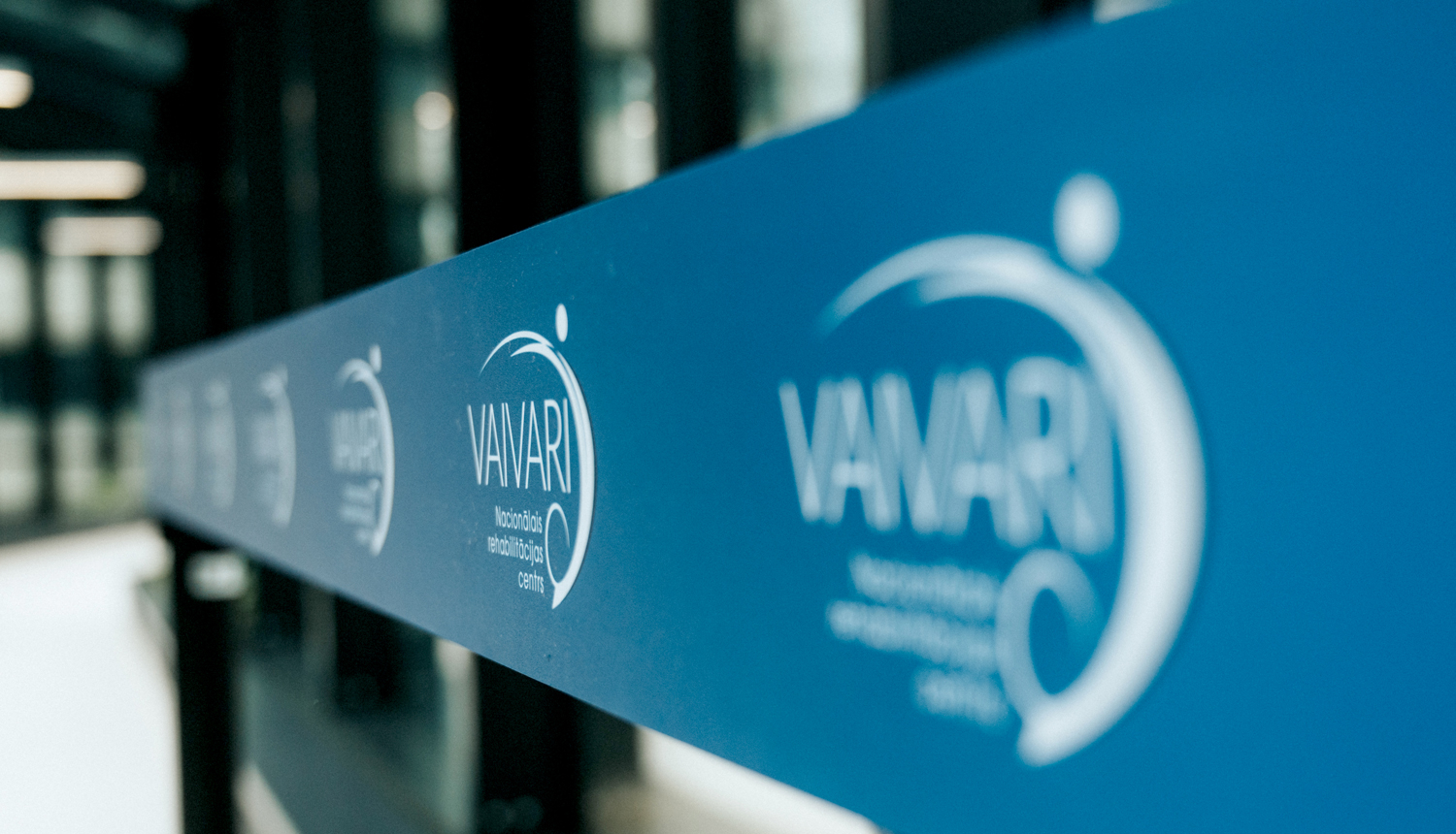 Zila stikla vai sienas virsma ar atkārtotu baltu “VAVARI” logotipu, fotografēta perspektīvā garā gaitenī.