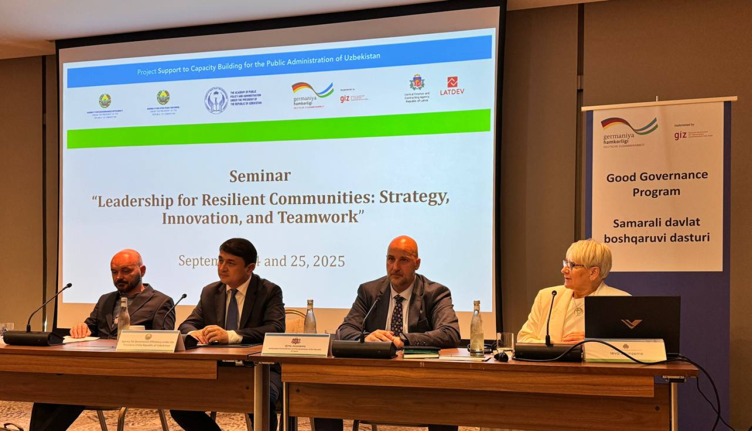 Seminārs “Līderība noturīgām kopienām: stratēģija, inovācijas un komandas darbs”, kas notika 2025. gada 24. un 25. septembrī. Attēlā redzami četri cilvēki, kas sēž pie garas galda ar mikrofonu priekšā. Fonā redzams liels ekrāns ar semināra nosaukumu, datumu un organizāciju logotipiem. Labajā pusē novietots plakāts ar uzrakstu “Good Governance Program” angļu un citā valodā. Telpa ir konferenču zāle ar paklāju un profesionālu iekārtojumu