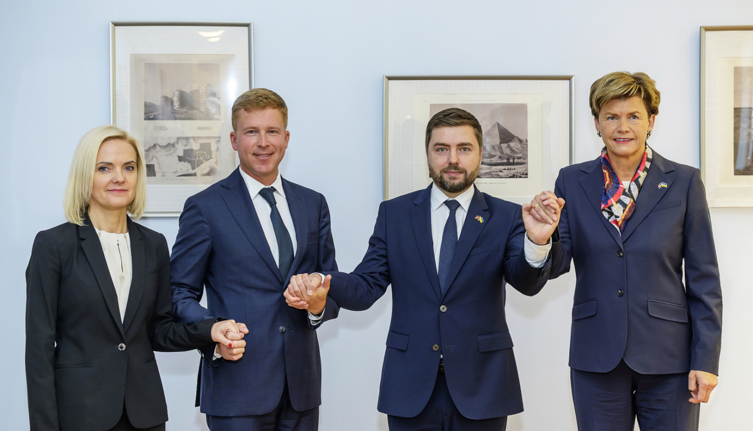 Četri cilvēki formālā biznesa apģērbā stāv rindā, sadevušies rokās, oficiāla pasākuma laikā pie sienas ar ierāmētiem attēliem fonā.