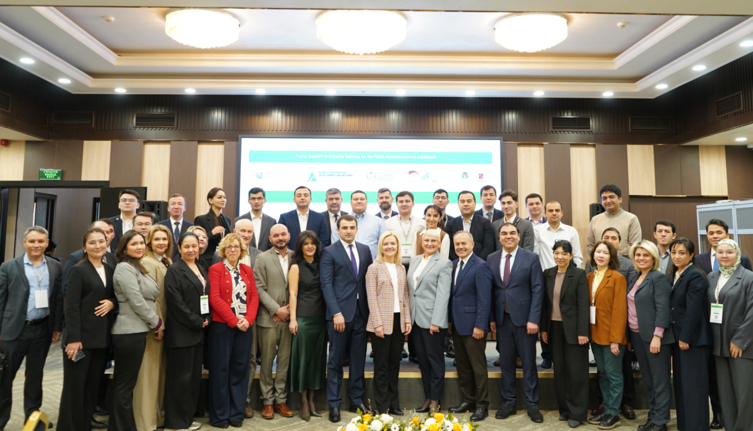 Liela grupa ar projekta dalībniekiem un Latvijas ekspertiem uz skatuves Taškentā, noslēdzot stratēģiskās plānošanas apmācību semināru. Fonā redzams ekrāns ar projekta nosaukumu "Project Support to Capacity Building for the Public Administration of Uzbekistan".