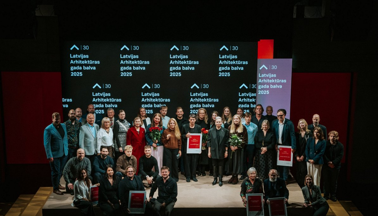 Grupas fotogrāfija ar cilvēkiem, kas stāv uz skatuves un tur rokās balvas. Fonā redzams ekrāns ar uzrakstu "Latvijas Arhitektūras gada balva 2025"