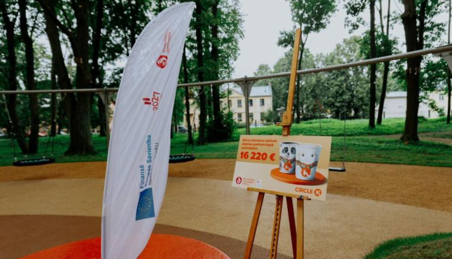 Āra teritorija ar rotaļu laukumu fonā. Priekšplānā redzama balta karogs ar tekstu un logotipiem, kā arī molberts ar plakātu, uz kura attēloti divi bundžu attēli un teksts: "Bērnu pilsētas aktivitāšu centra izveidei piešķirts līdzfinansējums 16 220 €".