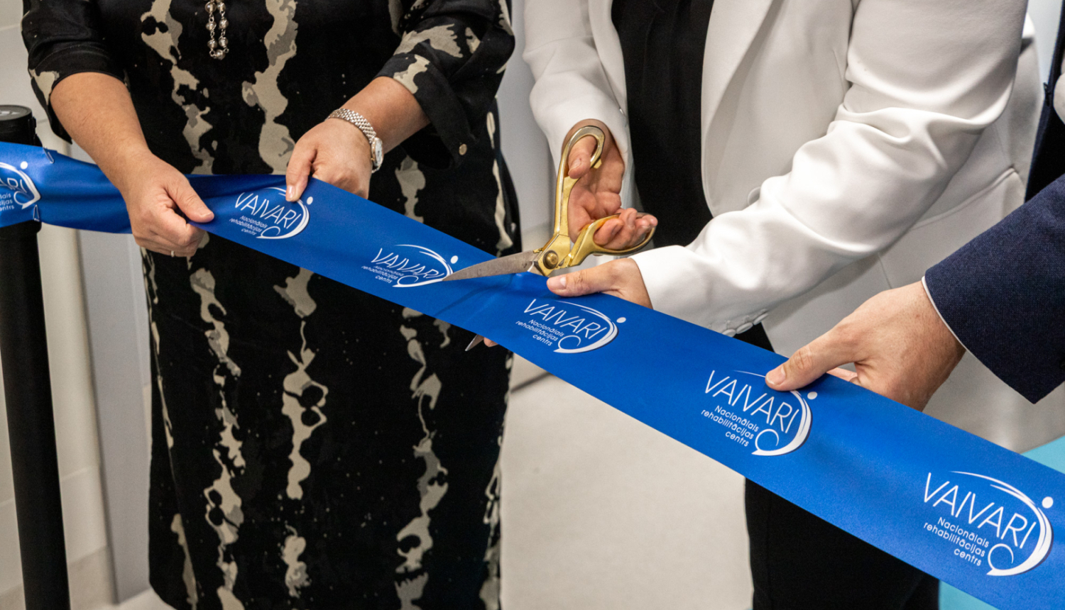 Trīs cilvēki piedalās lentes griešanas ceremonijā pie VAIVARI Nacionālā rehabilitācijas centra. Viens no viņiem ar lielām zelta šķērēm pārgriež zilu lenti ar centra nosaukumu, atzīmējot jaunas ēkas vai pakalpojuma atklāšanu.