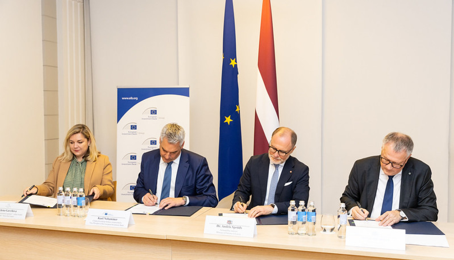 4 pārstāvji paraksta dokumentus pie galda, fonā redzamas Eiropas Savienības un Latvijas karogi, kā arī Eiropas Investīciju bankas (EIB) baneris ar vietni www.eib.org. Attēls no oficiālas parakstīšanas ceremonijas.