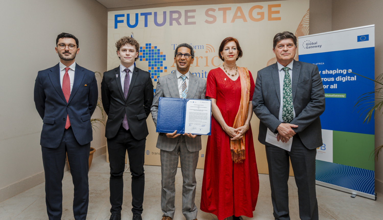 Pieci cilvēki stāv kopā pie sienas ar uzrakstu “Future Stage” un “Transform Africa Summit 2025”, viens tur atvērtu dokumentu mapē. Labajā pusē redzams plakāts ar Eiropas Savienības logotipu un tekstu par digitālās nākotnes veidošanu