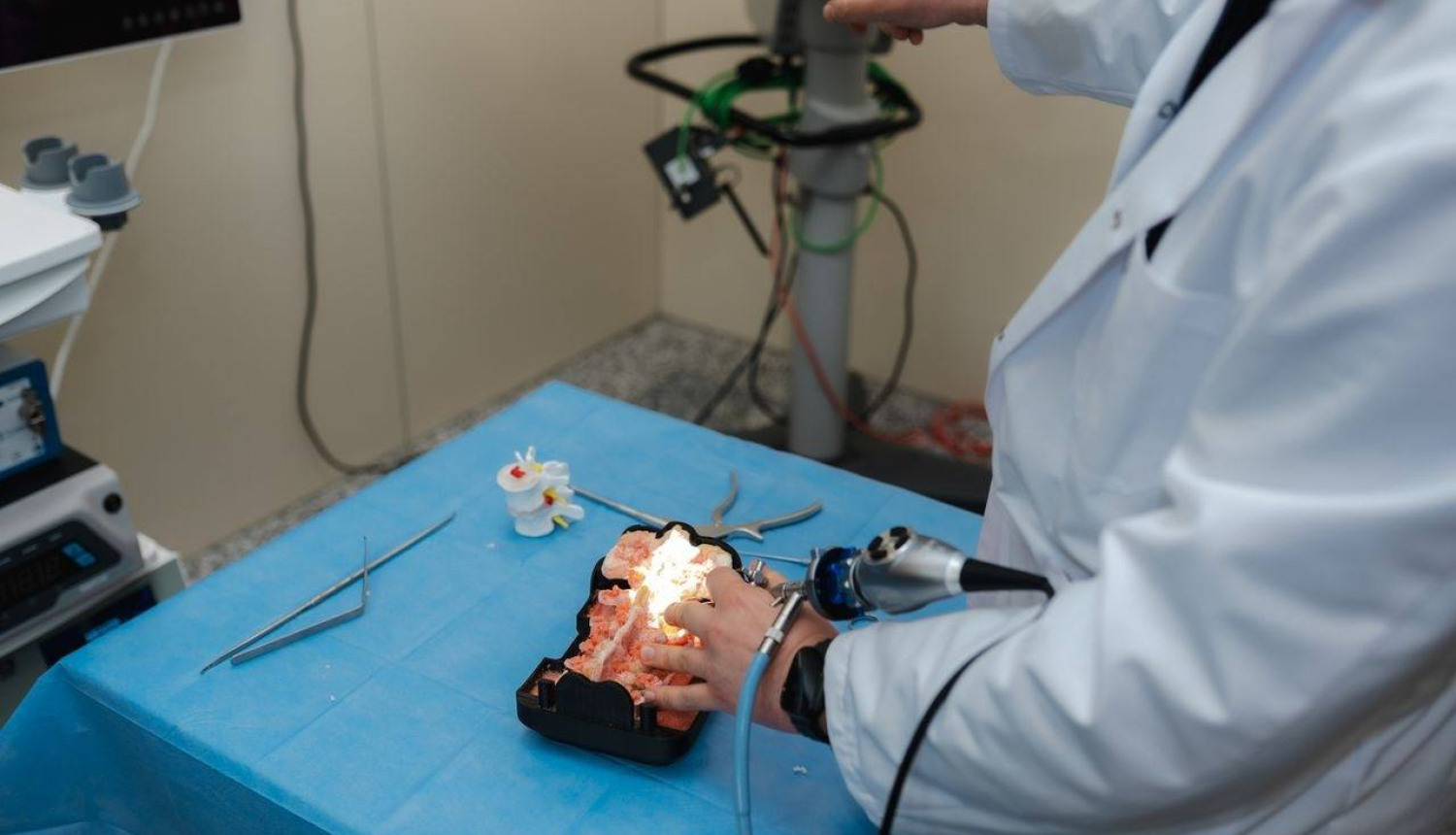 Persona baltā halātā veic manipulācijas ar medicīnisku vai anatomisku galvas/galvas smadzeņu modeli, kas novietots uz zilas ķirurģiskās drānas. Tiek izmantoti medicīnas instrumenti, tostarp endoskops vai tam līdzīga ierīce. Apkārt redzami dažādi rīki un aprīkojums, norādot uz medicīnas laboratoriju vai apmācību telpu, kur notiek praktiska procedūra vai mācību sesija.