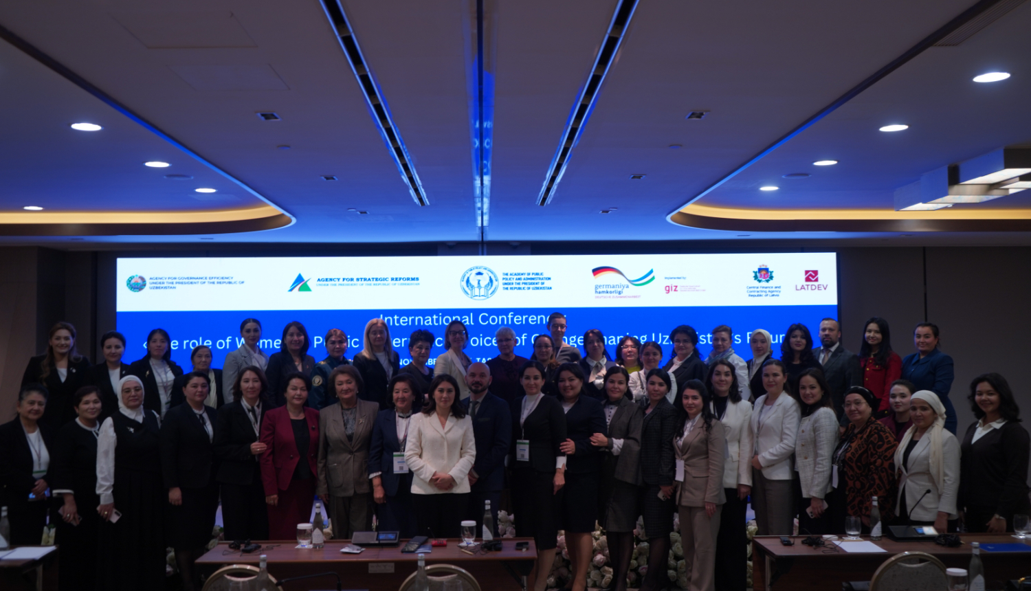 Grupas foto no starptautiskās konferences par dzimumlīdztiesību valsts pārvaldē Taškentā; dalībnieki pozē kopā konferenču zālē, fonā redzams liels ekrāns ar konferences nosaukumu un partneru logotipiem.