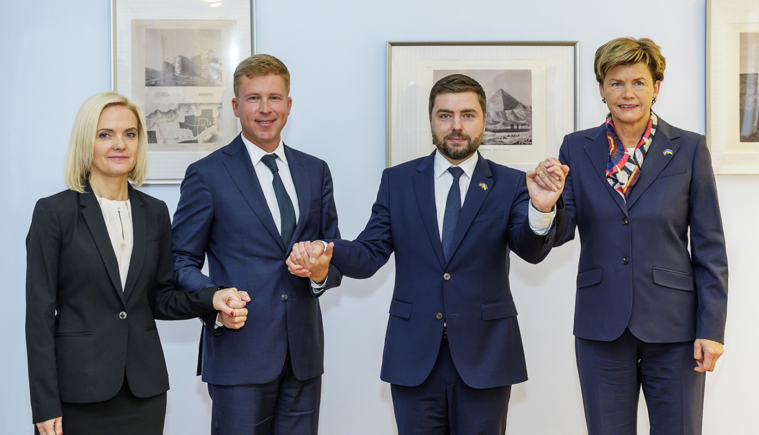 Četri cilvēki formālā biznesa apģērbā stāv rindā, sadevušies rokās, oficiāla pasākuma laikā pie sienas ar ierāmētiem attēliem fonā.