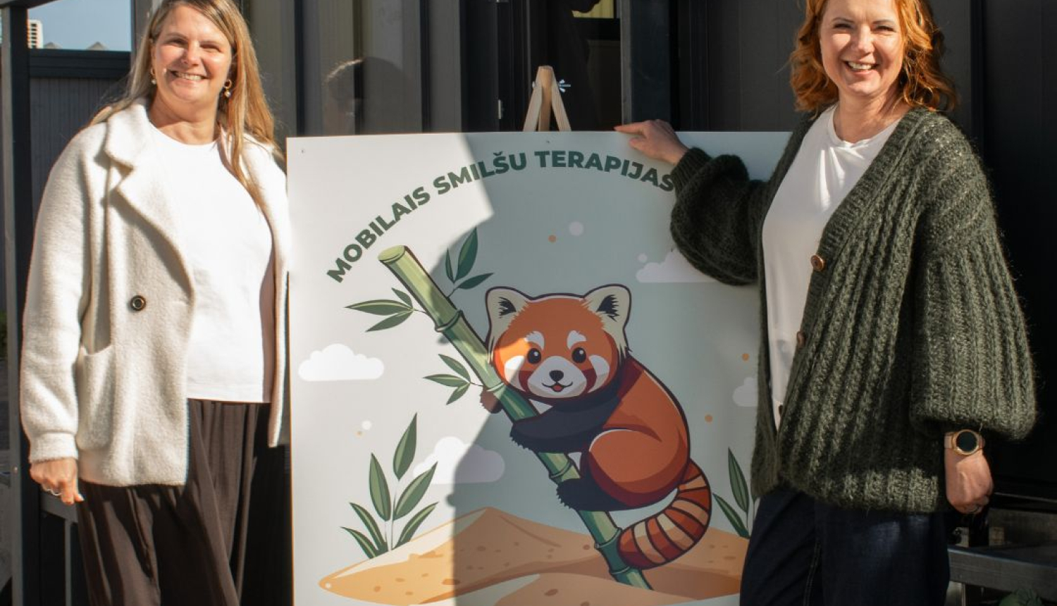 Divi cilvēki stāv pie plakāta ar ilustrāciju, kurā redzama sarkana panda ar bambusu. Uz plakāta rakstīts 'Mobilais smilšu terapijas'. Fonā redzami augi un smilšu kāpas, radot dabas un terapijas noskaņu